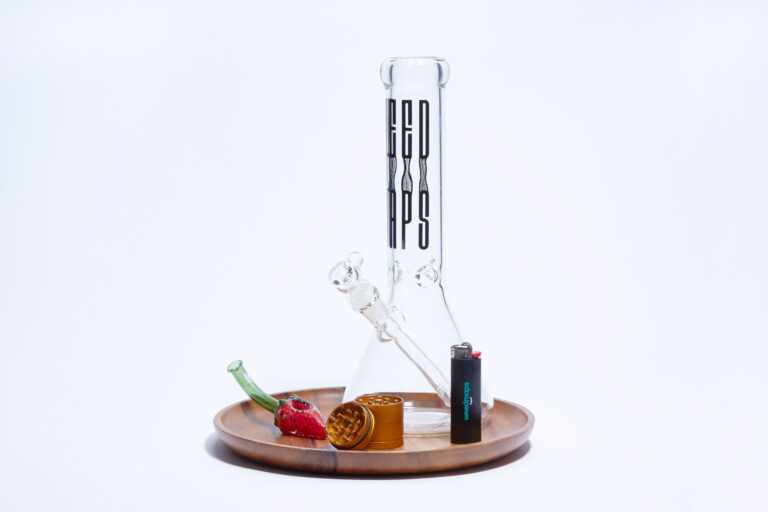 Bong, pipe, grinder, and lighter on wood platter on white background.
