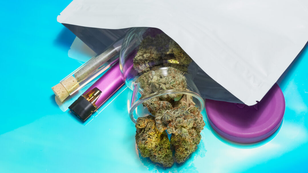 Various cannabis products spilling out of small white bag on teal background.