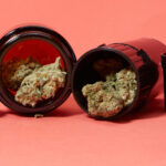 Cannabis nugs coming out of two black storage jars on pink background.