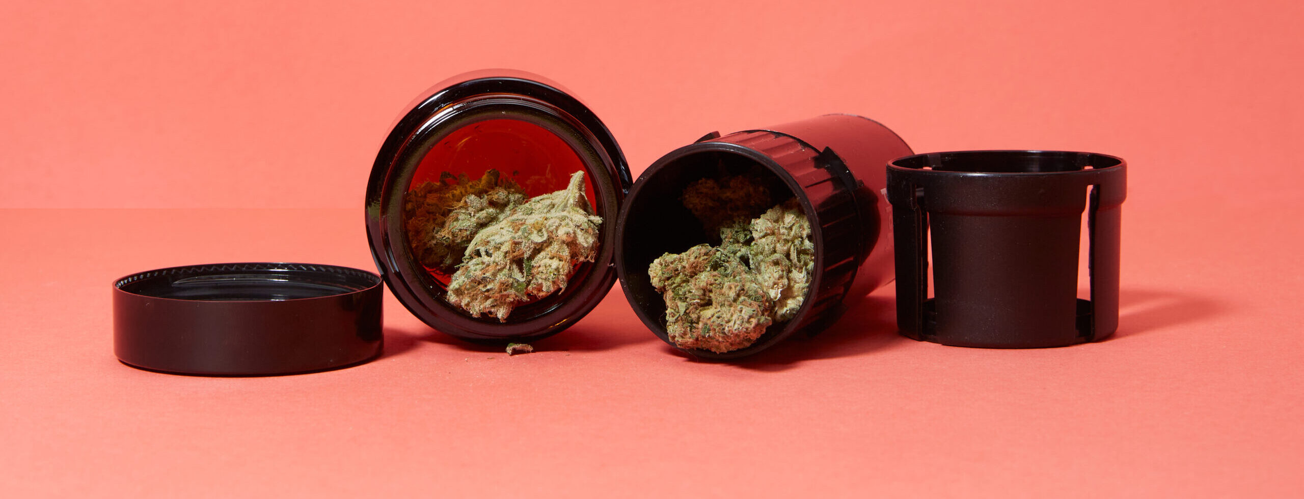 Cannabis nugs coming out of two black storage jars on pink background.
