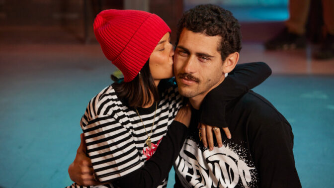 Young couple embracing intimately by a pool, woman in red beanie and striped shirt kissing man's cheek