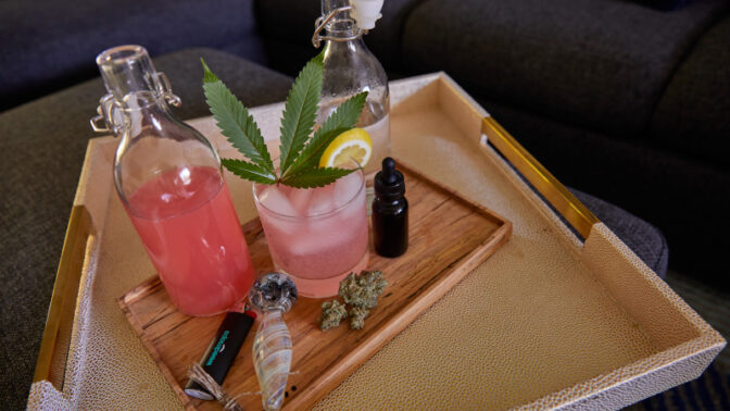 Cannabis-infused pink cocktail with marijuana leaf garnish on wooden serving tray alongside glass bottles, dropper bottle, and cannabis buds