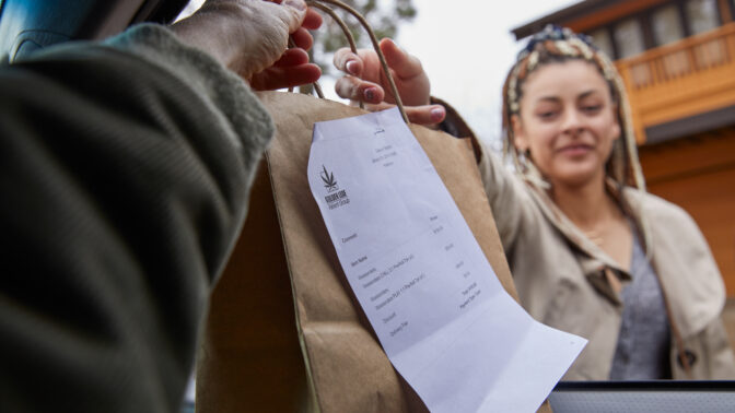A delivery driver handing a brown paper bag weed delivery to a woman outside the car